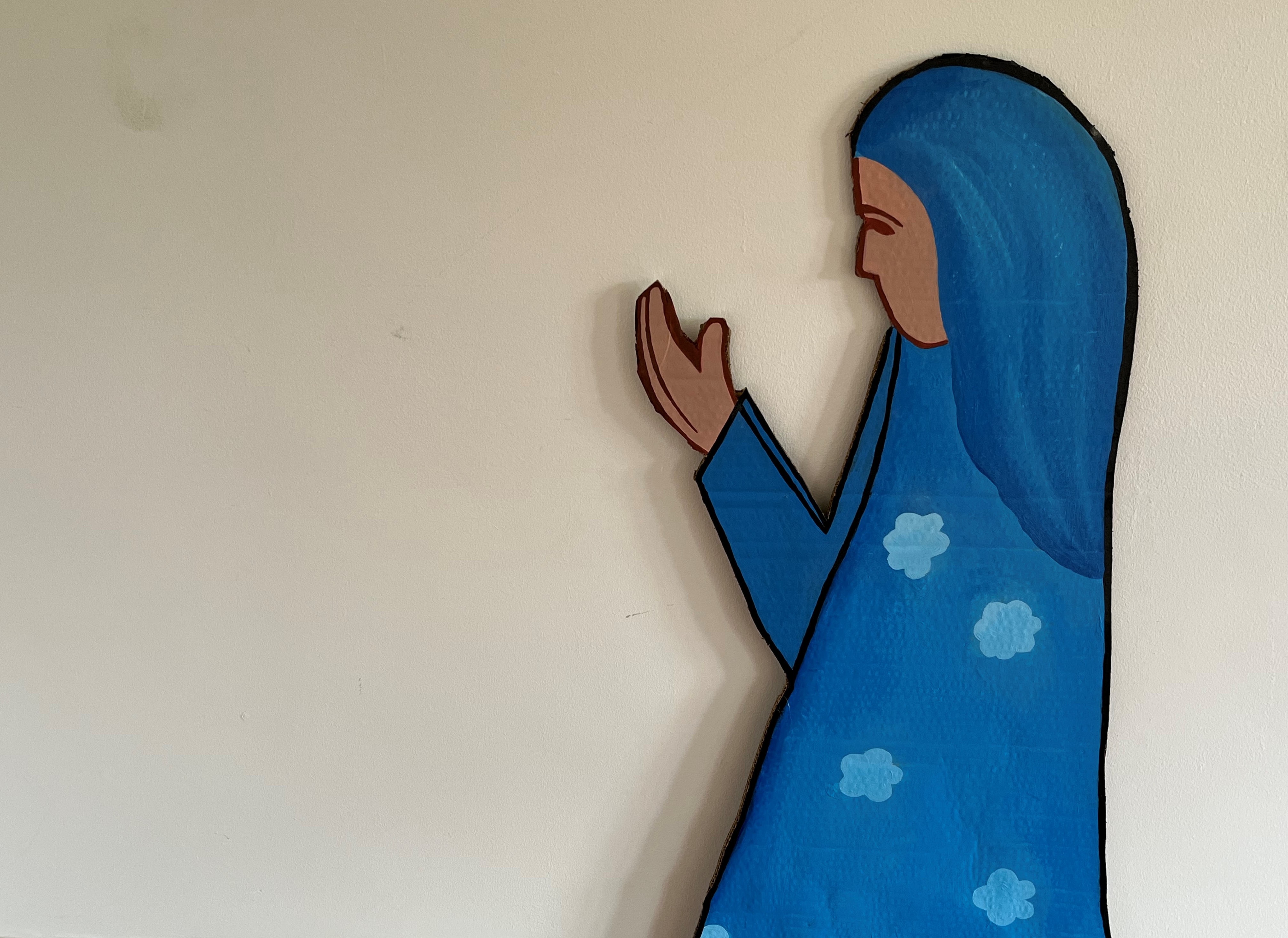 A woman wearing a blue veil and dress looks to her hands in prayer, her dress has light blue blossoms on it